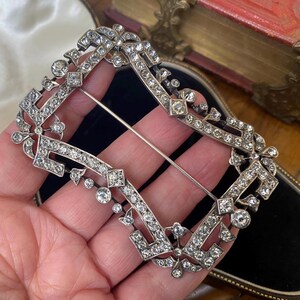 May include: An ornate silver-toned brooch with a rectangular shape, encrusted with numerous clear, sparkling gemstones. The brooch features a pin on the back and intricate floral and geometric designs. The jewelry is displayed against a dark background.