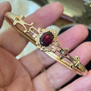 May include: A gold-toned hinged bangle bracelet featuring an oval-cut deep red garnet gemstone at the centre. The metalwork displays intricate scrollwork and filigree detailing surrounding the stone, with a smooth, polished band on the sides.