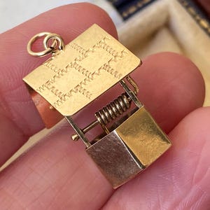 May include: A gold-toned metal charm shaped like a miniature wishing well, featuring a rectangular roof with a textured zigzag pattern, a central horizontal bar with a coiled spring mechanism, and a small bucket suspended at the base.