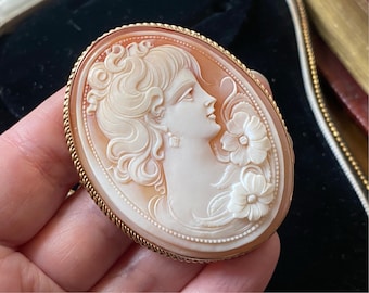 Könnte beinhalten: Eine goldfarbene Kameenbrosche mit einem geschnitzten Muschel-Kamee eines Frauenprofils mit detaillierten Haaren und floralen Akzenten.