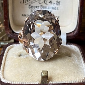 Puede incluir: Un anillo de piedra preciosa de talla ovalada con una superficie facetada, engastado en una banda de oro. El anillo se exhibe en un joyero vintage con un interior color crema y un exterior marrón oscuro. La piedra preciosa tiene un tono marrón claro.