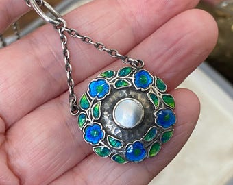 Puede incluir: Un colgante circular de plata con una incrustación central de nácar blanco rodeada por una textura de metal martillado. El borde exterior está decorado con flores y hojas de esmalte azul y verde, suspendido de una cadena de plata.