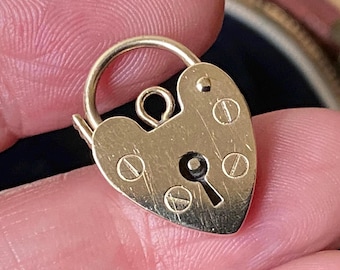 May include: A gold-colored heart-shaped padlock with a curved top and keyhole. The padlock has four small screws and a small loop at the top. The padlock is held in a person's hand.
