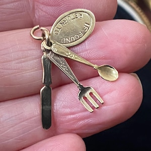 Op de afbeelding: Een gouden bedel bestaande uit een miniatuurmes, vork en lepel bevestigd aan een klein ovaal plaatje. Het plaatje is voorzien van de tekst IF FOUND RETURN TO. De drie keukengerei-onderdelen zijn verbonden door een kleine ring aan een oogje.