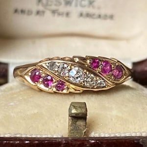 Może przedstawiać: Złoty pierścionek o liściastym wzorze, wysadzany przezroczystymi i różowymi kamieniami szlachetnymi. Pierścionek jest prezentowany wewnątrz zabytkowego pudełka na biżuterię z kremowym wnętrzem. Kamienie szlachetne pierścionka są rozmieszczone symetrycznie.