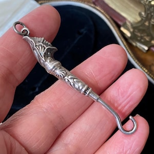 May include: A silver antique button hook with decorative floral and shell-like patterns. The hook has a curved end and a loop at the top. The item is held in a hand, indicating its small size and delicate nature.