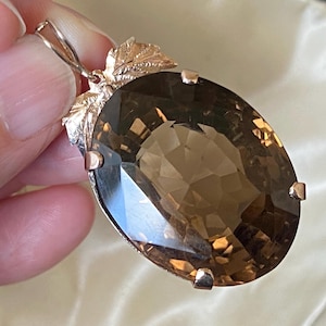 Puede incluir: Colgante de cuarzo ahumado de corte ovalado con engaste dorado. El colgante presenta una piedra preciosa facetada y está adornado con detalles decorativos de hojas. El engaste incluye una argolla para colgar el colgante.