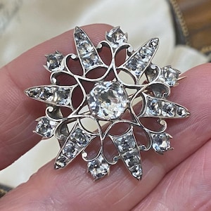 Könnte beinhalten: Eine silberfarbene Brosche in Sternform mit einem großen, facettierten, klaren Stein in der Mitte, umgeben von kleineren klaren Steinen, die in einen strahlenförmigen Metallrahmen mit filigranen Schnörkeln zwischen den Sternspitzen gefasst sind.