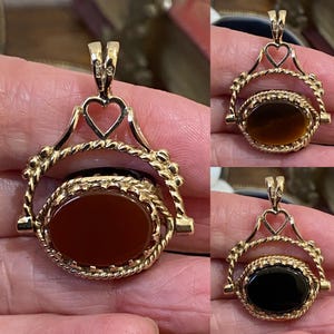 May include: A gold swivel pendant featuring an oval-shaped stone that rotates within a decorative frame. The frame includes a heart-shaped bail and twisted metal detailing. The pendant displays three different colored stones: reddish-brown, brown, and black.