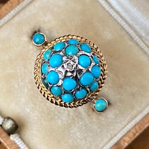 Puede incluir: Un anillo de oro con una piedra redonda de turquesa rodeada de piedras de turquesa más pequeñas. El anillo está engastado en una banda de oro y tiene un diseño delicado.