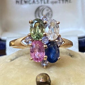 Puede incluir: Un anillo de oro con un grupo de piedras preciosas de forma ovalada. Las piedras son verdes, rosas, azules y blancas.