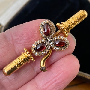 以下が含まれることがあります： 3つの楕円形の深い赤色の宝石と小さな透明な石が特徴の、金色のアンティークブローチ。ブローチは、湾曲したステムと2つの円筒形要素を備えた装飾的なデザインです。