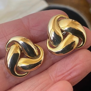 May include: A pair of polished gold-tone metal earrings featuring a three-dimensional, interlocking knot design. Each earring has a smooth, reflective surface with curved, rounded segments that loop together to form a continuous, circular shape.