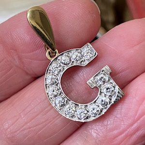 Puede incluir: Un colgante dorado con forma de la letra "G", incrustado con numerosas piedras preciosas claras y brillantes. El colgante tiene un pequeño lazo en la parte superior para una cadena. Las piedras están engastadas en metal plateado.