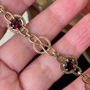 Puede incluir: Una pulsera de metal en tono dorado con un patrón repetitivo de eslabones circulares texturizados y conectores en forma de infinito. El diseño alterna entre estos eslabones circulares, engastados con piedras redondas de color rojo.