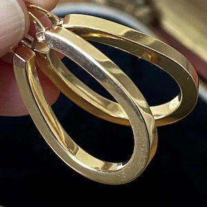 Puede incluir: Un par de pendientes de aro dorados. Los pendientes tienen forma rectangular y un acabado pulido. Los aros están sostenidos por un dedo. Los pendientes son un artículo de joyería clásico.