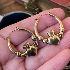 May include: A pair of gold-colored hoop earrings. Each earring features a Claddagh design, with a heart, crown, and hands. The earrings are displayed against a neutral background.