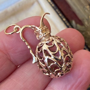 Puede incluir: Un colgante dorado con forma de jarrita. La jarrita tiene un diseño calado abierto, con asa y pico. El colgante está sujeto por la mano de una persona.