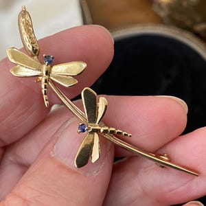 May include: A gold-toned brooch featuring two dragonfly figures perched on a curved stem. Each dragonfly has a small, round, dark blue gemstone set into its head. The metal surface is smooth and polished, with detailed wing textures.