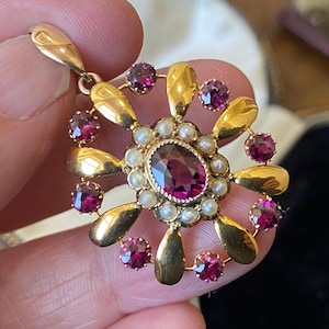 Puede incluir: Un colgante de oro con una piedra preciosa central ovalada de color rojo intenso, rodeada de pequeñas perlas. El colgante tiene un diseño floral con detalles dorados en forma de pétalos y piedras preciosas rojas redondas más pequeñas. Un lazo en la parte superior para colgar.