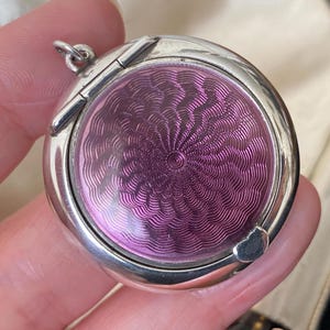 Puede incluir: Un medallón de plata con una tapa con bisagras y un diseño de esmalte rosa. El diseño es un patrón de círculos concéntricos en espiral.