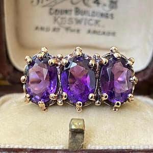 May include: A gold ring featuring three oval-cut purple amethyst gemstones set in a row. Each stone is held by multiple prongs, creating a continuous horizontal arrangement of deep violet gems against a metallic gold band structure.