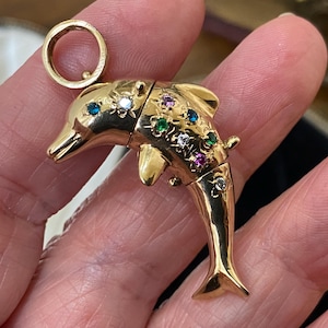Könnte beinhalten: Ein goldener Delphin-Anhänger mit einem aufklappbaren Körper, verziert mit bunten Edelsteinen. Der Anhänger hat oben eine kleine Schlaufe für eine Kette. Der Körper des Delphins ist mit Flossen und einem Schwanz detailliert.
