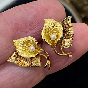May include: A pair of gold-toned metal earrings shaped like calla lilies with textured surfaces and a single small white pearl accent in the center of each flower. The earrings feature a detailed leaf design and curved stems, showcasing a metallic finish.