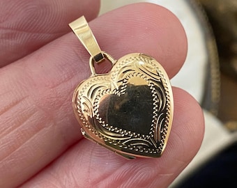 May include: A gold heart-shaped locket pendant featuring an engraved border with decorative scrollwork and a central smooth heart-shaped panel. The pendant includes a tapered bail at the top for attachment to a chain, showcasing a polished finish.