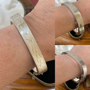 May include: A silver-toned hinged bangle bracelet featuring an engraved leaf pattern across the outer surface. The bracelet includes a secure clasp mechanism and a small safety chain attached to the side for added security while worn.