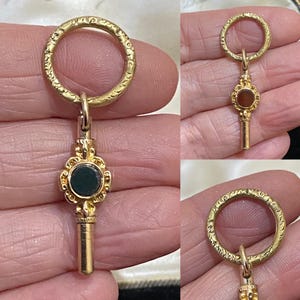 May include: A gold pocket watch key with a black stone set in a decorative frame. The key has a round loop at the top and a long, thin shaft.