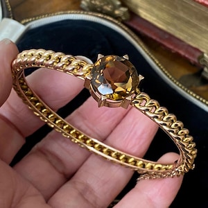 Puede incluir: Un brazalete dorado con una gran gema redonda de color ámbar en el centro. El brazalete presenta un diseño de eslabones de cadena y se sostiene en la mano. El fondo es una caja oscura forrada de terciopelo.