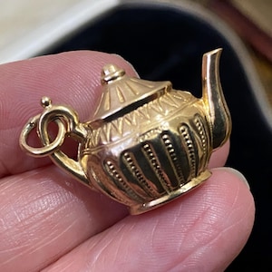 May include: A small gold-toned metal charm shaped like a miniature teapot. The charm features a detailed lid with radial lines, a curved spout, and a body decorated with vertical ribbed patterns. A small circular jump ring is attached to the handle.