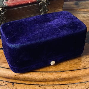 Puede incluir: Un joyero rectangular de terciopelo morado oscuro con cierre de botón nacarado. La caja está colocada sobre una superficie de madera con un diseño decorativo. Una parte de un libro antiguo es visible en el fondo.