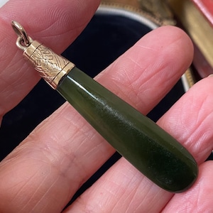 Puede incluir: Un colgante en forma de lágrima con una piedra verde oscuro y detalles metálicos dorados. La parte superior del colgante tiene un diseño de hojas. El colgante está sostenido en una mano.