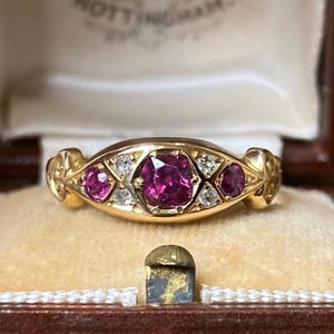 May include: A gold ring featuring a central cushion-cut deep pink gemstone flanked by two smaller round-cut pink gemstones and four tiny clear accent stones. The metal band has a polished finish with decorative engraved details on the sides.