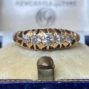 May include: A gold ring featuring five round faceted clear stones set in a row. The band has a decorative carved pattern around each stone setting. The ring is displayed in a blue velvet-lined box with the text NEWCASTLE UPON TYNE visible.