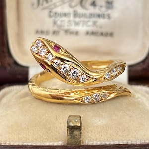 May include: A gold snake ring featuring a wrap-around design with a textured body. The head and tail are set with clear round-cut stones, and the eyes are accented with small red stones. The ring is displayed in a vintage-style jewelry box.