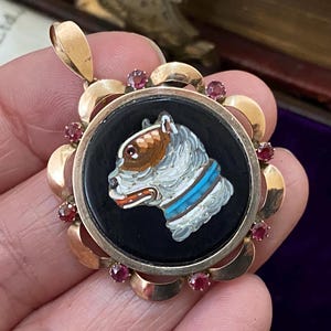 May include: A gold pendant featuring a central black circular inlay with a micro-mosaic dog profile in white, brown, and blue. The frame is decorated with scalloped gold edges and six small round red gemstones set around the perimeter.