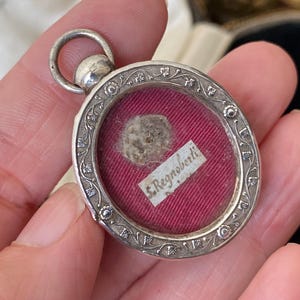 Pode incluir: Um relicário de prata com um design floral e um inserto de vidro que contém um pequeno pedaço de tecido e uma nota manuscrita que diz "St. Regnoberti".