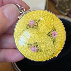 May include: A round, yellow pendant with a silver clasp. The pendant features a floral design with pink roses and green leaves. The background has a textured pattern.