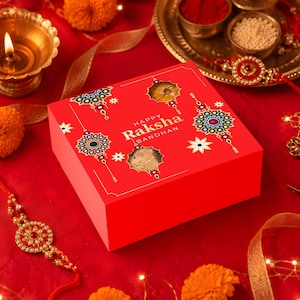 Può includere: Una scatola regalo quadrata rossa con la scritta Happy Raksha Bandhan stampata in bianco. La scatola presenta motivi circolari decorativi in verde, blu e oro, e due aperture circolari che rivelano il cibo contenuto all'interno.