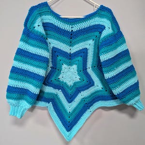 May include: A handmade, star-shaped crochet sweater in shades of blue and turquoise. The sweater features a large star design in the center, with striped sleeves and a unique, asymmetrical hemline. The sweater is displayed on a white hanger against a gray background.