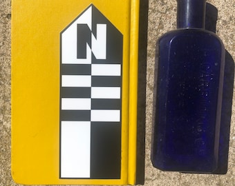 Könnte beinhalten: Ein gelbes Notizbuch mit einem schwarz-weißen Aufkleber, der einen stilisierten Buchstaben "N" und ein Schachbrettmuster zeigt. Das Notizbuch liegt neben einer kobaltblauen Glasflasche.