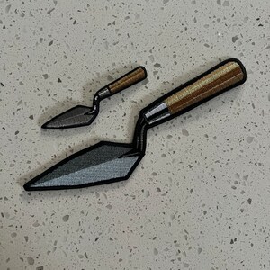 Puede incluir: Dos llanas, una peque&ntilde;a y una grande, con mangos de madera marr&oacute;n y hojas de metal gris. Las llanas est&aacute;n sobre una superficie blanca moteada.