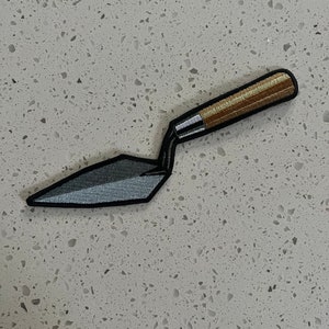 Könnte beinhalten: Ein gestickter Aufnäher, der eine Kelle mit einem Holzstiel in Grau- und Brauntönen zeigt.