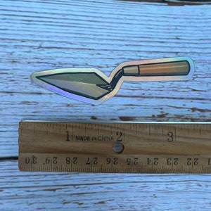 Peut inclure: Un autocollant iridescent argenté et brun d'une truelle, un outil utilisé pour étaler du mortier ou du plâtre.