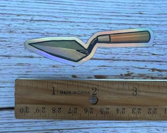 May include: A silver and brown iridescent sticker of a trowel, a tool used for spreading mortar or plaster.