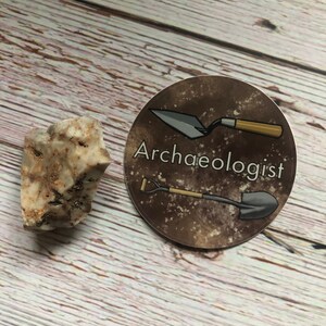 Può includere: Un adesivo rotondo con sfondo marrone e testo bianco che dice "Archeologo". L'adesivo presenta una pala nera e una cazzuola con manico in legno.