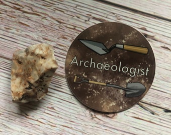 May include: A round sticker with a brown background and white text that says "Archaeologist". The sticker features a black shovel and a trowel with a wooden handle.